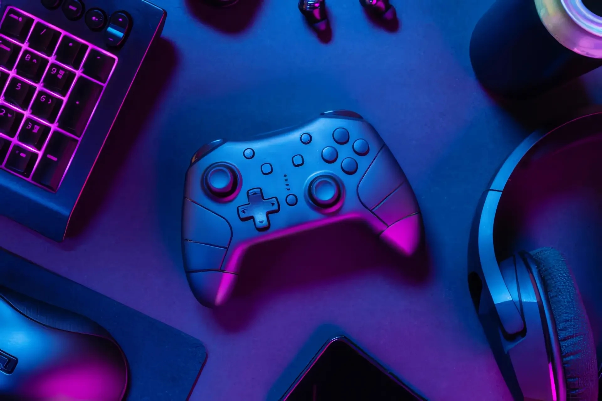 An overhead view of a game controller on a desk, surrounded by a keyboard, mouse, headset, and phone, lit in purple and blue.